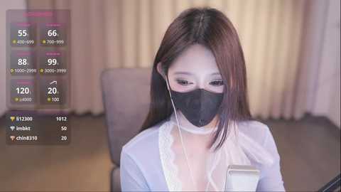 Media: Video of an East Asian woman with long brown hair, wearing a black face mask and a light blue shirt, seated in an office setting.