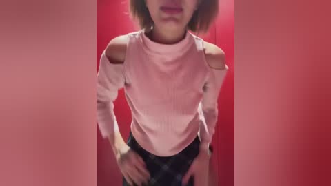 Media: Video of a slender, light-skinned woman with short brown hair, wearing a pale pink, sleeveless top with shoulder cutouts and black panties, against a bright red backdrop.