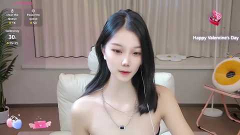 Media: A video of a slender Asian woman with long black hair, wearing a black strapless top and a necklace, sitting in a modern living room with beige curtains, a white chair, and a pink table lamp.