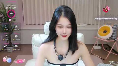 Media: Video of an East Asian woman with long black hair, wearing a strapless black top, sitting in a white leather chair. Background features a pink heart-shaped fan, a potted plant, and a Valentine's Day message.