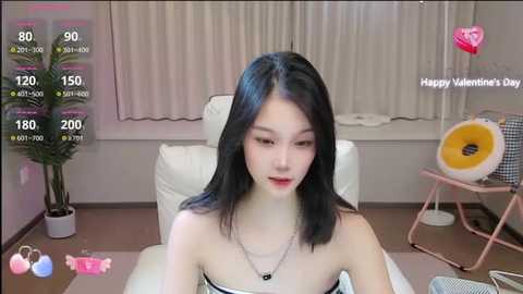 Media: A video of an East Asian woman with straight black hair and fair skin, wearing a strapless black dress, sitting on a white couch in a modern, minimalist room.