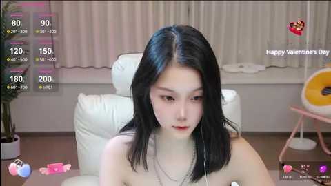 Media: Video of an Asian woman with straight black hair, light skin, wearing a black top, sitting on a white leather chair in a room with white curtains, pink heart-shaped objects, and a pink lamp.