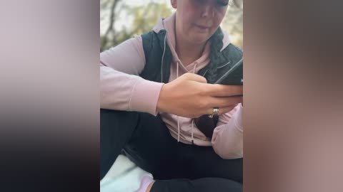 Media: Video of a young woman with light skin, wearing a pink hoodie, black vest, and dark pants, sitting outdoors, texting, blurred background.