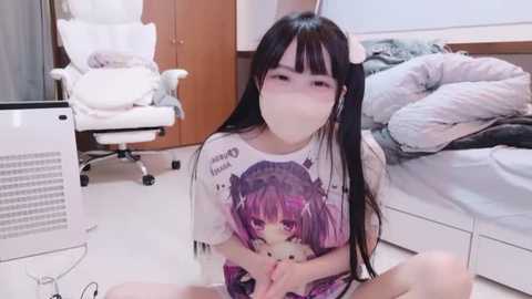 Media: Video of a young Asian woman with long black hair, wearing a white T-shirt with a purple anime character, sitting on the floor in a tidy, minimalist bedroom.