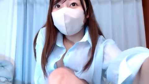 Media: Video of an Asian woman with long brown hair, wearing a white surgical mask and light blue button-up shirt, sitting on a bed. The background features light blue curtains and a pillow.