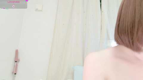 Media: Video of a woman's bare back and auburn bob haircut in a white-tiled bathroom. A pink hairbrush is on the left wall, and a white curtain covers a window in the background.