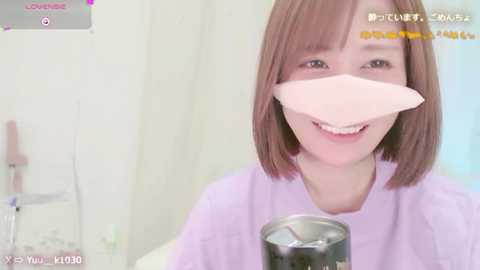 Media: A video of a young woman with short brown hair, wearing a light purple top and a surgical mask, smiling while holding a black metal cup. The background features a white wall with medical equipment, indicating a hospital setting.