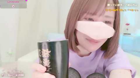 Media: Video of a young, light-skinned woman with shoulder-length brown hair, wearing a purple t-shirt and a black face mask. She holds a black ceramic mug with floral design. Background is a blurred, white room with a hint of a bed.