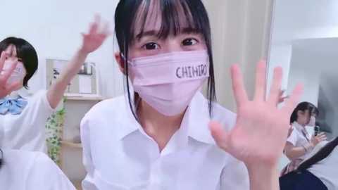 Media: Video of a young Asian woman with straight black hair, wearing a white blouse, pink mask, and blue bow, waving enthusiastically in a brightly lit, modern room.