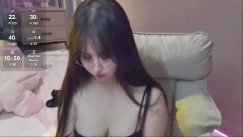 Media: A video of a young Asian woman with long black hair, wearing a black bra, sitting on a beige sofa. The background shows a dimly lit room with a pink wall and a wooden dresser.