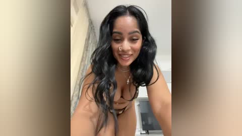 Media: Video of a smiling, light-skinned woman with long, wavy black hair and a nose ring, wearing a brown bra, leaning forward in a bathroom with beige walls.