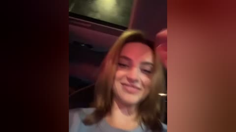 Media: Video of a young Caucasian woman with shoulder-length blonde hair, smiling and looking down, seated in a dimly lit car interior.