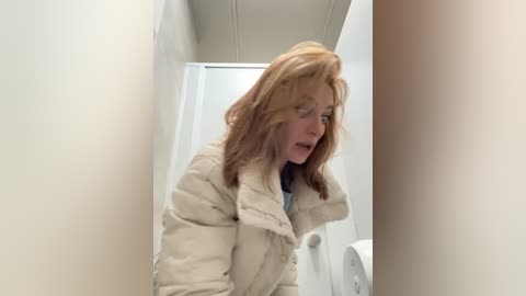 Media: Video of a young woman with light brown hair, wearing a puffy white jacket, looking downward in a narrow, dimly lit bathroom.