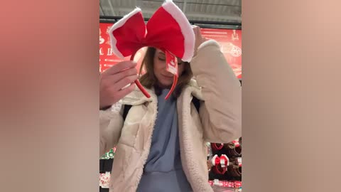 Media: Video of a young woman wearing a festive red and white Santa hat with a large bow, a blue sweater, and a beige fur-trimmed jacket, standing in a holiday-themed store with red and white decorations and Christmas ornaments in the background.