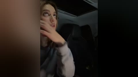 Media: A dimly-lit video of a young woman with light skin and shoulder-length brown hair, wearing a white shirt, peering out from the window of a dark car, fingers on her cheek.