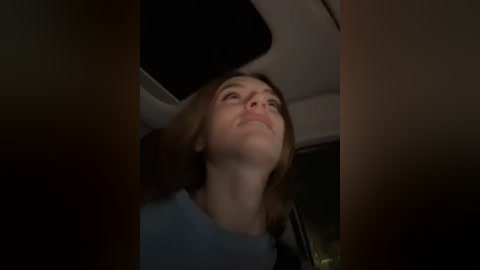 Media: A video of a young woman with light skin and shoulder-length brown hair, wearing a light blue shirt, looking up with a contemplative expression. The background shows the dim interior of a vehicle.