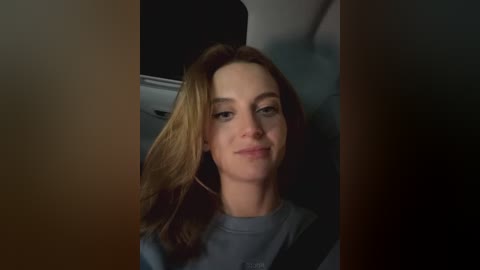 Media: Video of a smiling woman with long, wavy, light brown hair, wearing a gray shirt, seated in a car with dark, dim lighting.