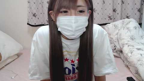 Media: A video of a young Asian woman with long straight brown hair, wearing a white mask and t-shirt, in a dimly lit room with white floral curtains and a messy bed.