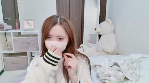 Media: Video of an Asian woman with light skin and long, straight brown hair, wearing a white mask, holding a white teddy bear in a white and beige room.