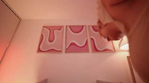 Media: A video of a blonde woman with short hair, wearing a white tank top, leaning forward in a dimly lit room. The background features a wall with three abstract paintings of wavy, pink and white patterns.