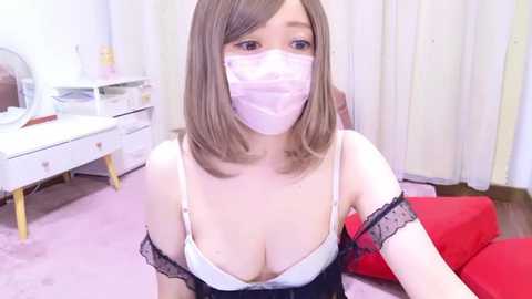 Media: Video of an Asian woman with shoulder-length brown hair, wearing a pink surgical mask, black lace-trimmed bra, and white camisole, sitting on a red cushion in a pink-carpeted room with white curtains and a white desk.