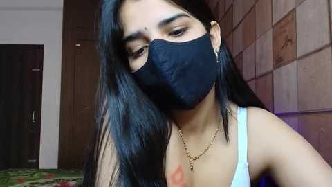 Media: Video of a young woman with long black hair, wearing a black mask, light blue tank top, and a gold necklace, standing indoors against a tiled wall.