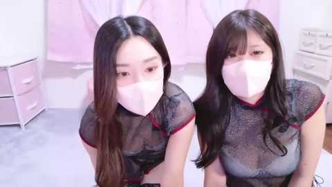 Media: Video of two young Asian women with long black hair, wearing white masks, black mesh tops, and red trim, sitting on the floor in a pale pink room with pastel furniture.