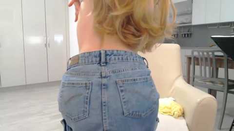 Media: A video of a blonde woman's back in tight blue jeans, standing in a modern kitchen with white cabinets, a cream chair, and a black floor lamp.