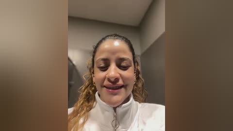 Media: Video of a smiling Latina woman with curly brown hair, wearing a white lab coat, standing indoors with beige walls.