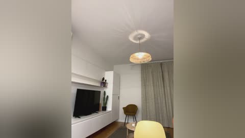 Media: Video of a minimalist living room with white walls, a flat-screen TV mounted on a white wall, a brown chair, a hanging pendant light, and a grey curtain.