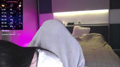 Media: A video of a person with long black hair wearing a white hooded sweater lying on a bed with a beige blanket. The room has a dark, modern ambiance with a digital clock display in the background.