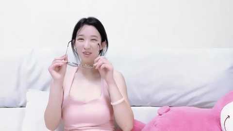 Media: Video of a young Asian woman with fair skin, wearing a light pink sleeveless dress, sitting on a white couch with a pink plush toy beside her.