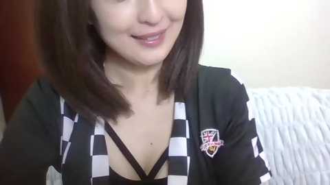 Media: Video of a smiling Asian woman with shoulder-length brown hair, wearing a black and white checkered cardigan over a black top with a crisscross neckline, and a sports team emblem on the left sleeve.