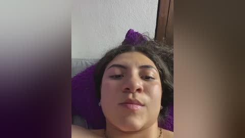 Media: A video of a young Latina woman with dark, curly hair and light skin, lying on a bed with a purple pillow, looking directly at the camera.