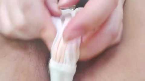 Media: Close-up video of a woman's shaved vulva, with light skin tone, using a white razor to shave. Her hands, with manicured nails, hold the razor.