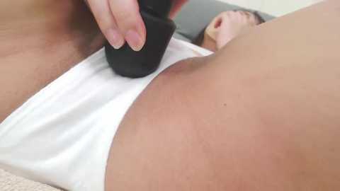 Media: Video of a close-up of a white woman's buttock, being massaged with a black vibrator. She wears white panties, lying on a grey bed, with a man's head visible in the background.