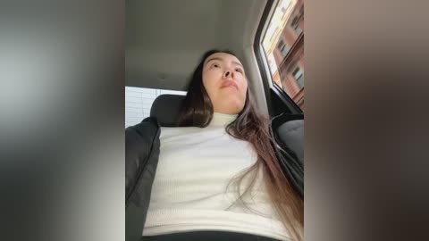 Media: Video of a woman with long, dark hair, wearing a white turtleneck, sitting in a car. She has a calm expression, looking out the window at a brick building.