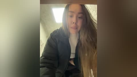 Media: A video of a young woman with long, wavy brown hair, wearing a black puffer jacket and white top, taken indoors, with a blurred background.