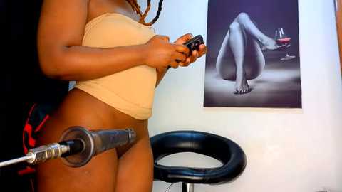 Media: Video of a woman with light brown skin wearing a beige strapless top and black bra, holding a black device connected to a metal tube. She stands beside a black leather chair in a minimalist, white room with a black-and-white artwork of a nude woman on the wall.