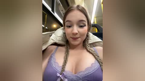 Media: A video of a fair-skinned, blonde woman with braided hair, wearing a lavender lace-trimmed camisole, smiling in a car with a beige jacket draped over the seat.