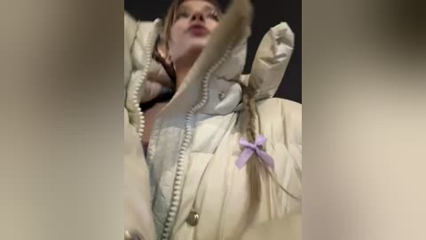 Media: Video of a young woman with light skin, wearing a cream-colored puffer jacket with a lavender bow, looking up with a slight smile.