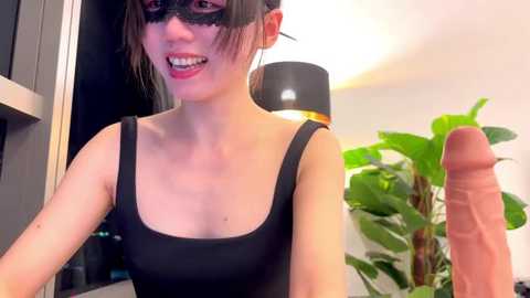 Media: Video of an Asian woman with fair skin and straight black hair, wearing a black mask and sleeveless black top, standing in a modern room with a green plant and a black lamp.