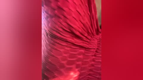 Media: Video of a woman's arm in a sleeveless, pleated, red dress with a hexagonal pattern, set against a solid red background. The fabric is smooth and the lighting is soft, emphasizing the dress's texture.