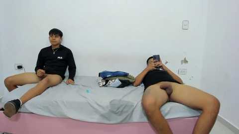 Media: Video of two young men, one wearing a black hoodie and no pants, sitting on a bed, the other in a cap, black shirt, and no pants, in a simple white-walled room.