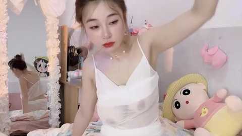 Media: A video of an Asian woman in a white camisole, with red lipstick, sitting on a bed adorned with plush toys, including a pink bear and yellow duck. The room is adorned with pastel decor.