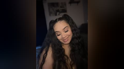 Media: Video of a smiling young woman with long, curly black hair styled in pigtails, wearing minimal makeup. She is in a dimly lit room with dark walls and a blurred, black-and-white framed photo on the wall.