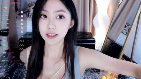 Media: Video of an East Asian woman with long black hair, fair skin, and a slim physique, wearing a gray tank top, taking a selfie in a bedroom with a bed, mirror, and soft lighting.