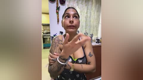 Media: Video of a Latina woman with dark skin and tattoos, wearing a black and white polka dot dress, making a peace sign in a kitchen with colorful hanging decorations.