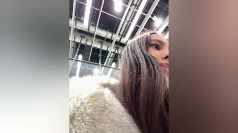 Media: Video of a woman with long, straight brown hair, wearing a white fur coat, standing indoors under a high ceiling with exposed beams and industrial lighting.