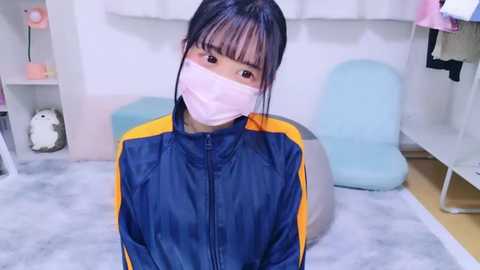 Media: Video of an East Asian woman with black hair, wearing a blue and yellow track jacket and pink face mask, sitting on a grey floor in a room with white walls and light furniture.